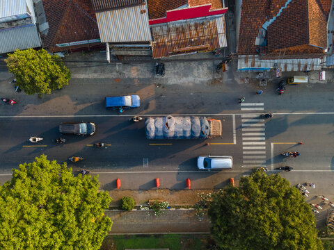 Aerial view of a busy road with cars, motorcycles, and a truck carrying a large load, flanked by lush green trees and buildings Cilongok, Central Java, Indonesia.