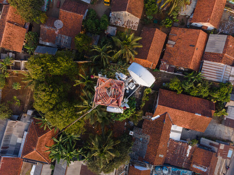 Aerial view of a telecommunications tower surrounded by residential houses with red-tiled roofs and lush palm trees in Cilongok, Central Java, Indonesia.
