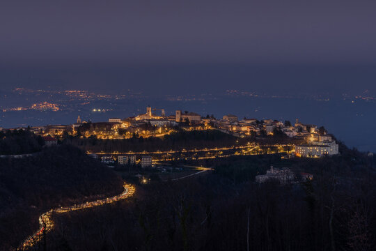 Aerial view of the illuminated hilltop town of Cingoli at night, with a winding road featuring car light trails leading up to the historic architecture in Cingoli, Marche, Italy.