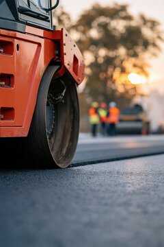 Low angle steamroller on new asphalt with construction crew in soft sunrise light for site banner and industrial background design