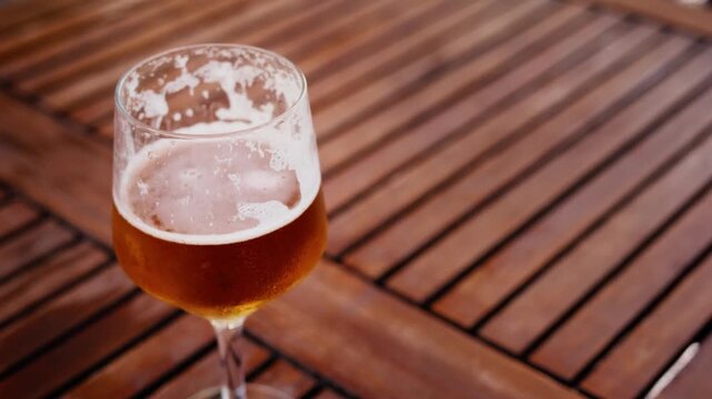 High-angle shot of a cold glass of amber beer with foam resting on a dark wooden slatted table outdoors in natural daylight.