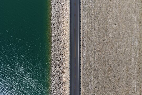 Aerial view of Horsetooth Reservoir dam featuring a paved road running along the crest, separating the deep blue water from the dry, rocky terrain in Larimer County, Colorado, United States.