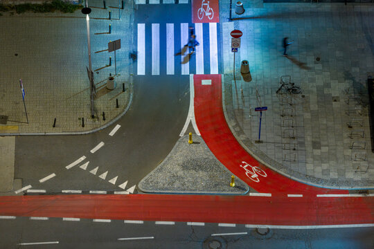 Aerial view of a city street intersection at night featuring a bright red bike lane, a pedestrian crosswalk with people walking, and urban road markings in Warsaw, Masovian Voivodeship, Poland.