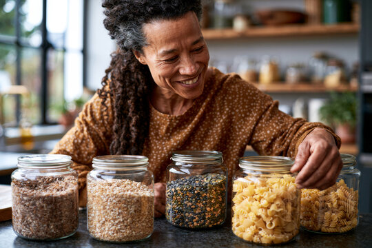 Smiling homeowner organizing reusable glass pantry containers filled with grains, legumes and pasta in a bright contemporary kitchen, warm window light