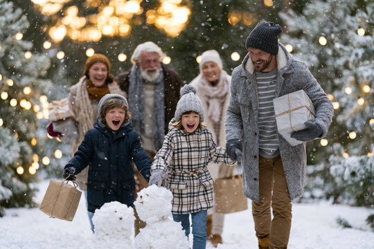 Contemporary arrangement of joyful multi-generational family celebrating the holiday season outdoors in a picturesque snow-covered landscape, building snowmen, laughing together, carrying wrapped