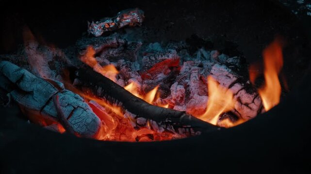 Close-up of glowing embers and burning firewood inside an outdoor fire pit at night.