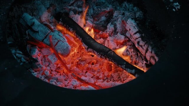 Close-up of glowing embers and burning firewood inside an outdoor fire pit at night.
