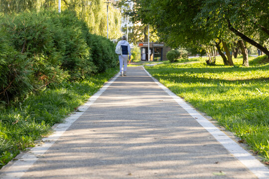 Asphalt walking path through green urban park in sunlight with pedestrian in distance