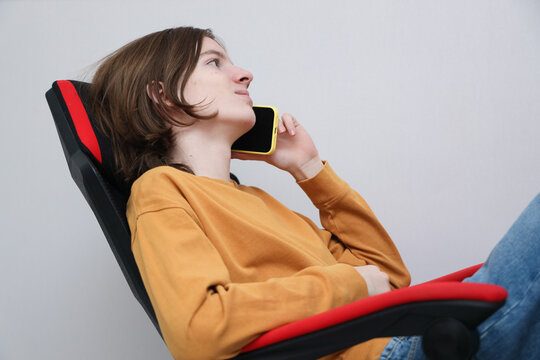 Young Person Reclining In Chair, Holding Smartphone To Ear, Wearing Mustard Sweater And Jeans, Shown In Calm Side Profile Against Plain Gray Background With Copy Space