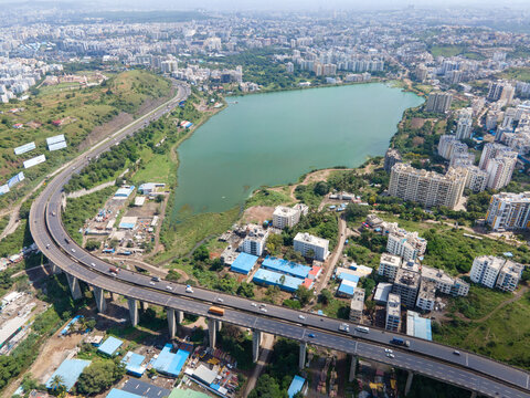 Aerial view of the Katraj highway flyover passing near the Katraj Lake, surrounded by urban residential buildings, green hills, and a clear sky in Pune, Maharashtra, India.
