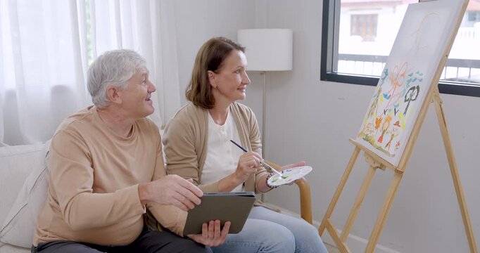 Senior woman paints colorful canvas while senior husband uses tablet and gestures nearby, sharing home art therapy supporting creativity, emotional wellness, companionship, active retirement.