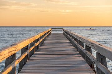 Fototapeta premium Luc-Sur-Mer, France - 08 08 2026: View of a wooden pier in front of the sea, a colorful sunrise and a fishing boat far away