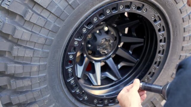 Person tightens lug nuts on rugged off-road tire. Hand holds red torque wrench with focus and care. Black wheel features deep treads and modern design. Perfect for vehicle maintenance, adventure