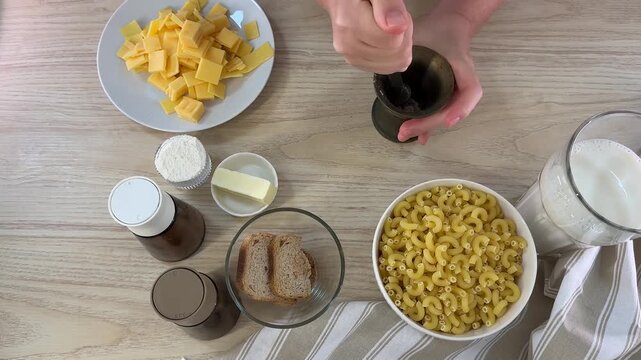 A person uses a mortar and pestle to grind ingredients on a wooden table, surrounded by bowls of macaroni, cheese slices, bread, and milk