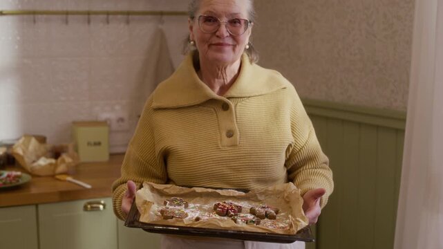 Portrait of smiling senior Caucasian woman holding baking sheet with decorated cookies in kitchen as camera tilts up. Clip fits holiday baking ads and family culinary promotions
