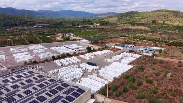 Overhead view of large plastic recycling plant with rows of white baled materials stored outdoors, solar panels on rooftop, surrounded by green hills and agricultural fields in Mediterranean landscape