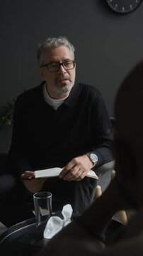 Vertical shot of middle aged Caucasian psychologist writing notes while listening to client during therapy session. Suitable for mental healthcare and medical projects