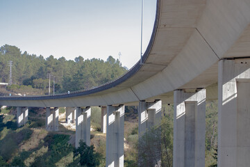Fototapeta premium Curved concrete highway viaduct with tall support pillars over a wooded valley, low angle view under the bridge with clear blue sky