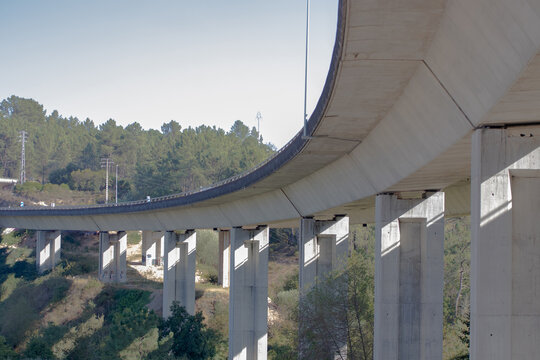 Curved concrete highway viaduct with tall support pillars over a wooded valley, low angle view under the bridge with clear blue sky