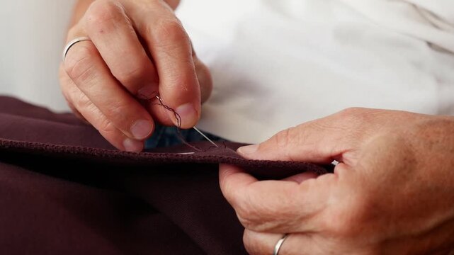 Macro close-up of senior woman sewing fabric by hand with needle and thread. Detailed clothing alteration and needlework process.