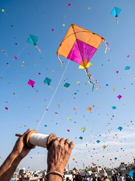 Hands holding a kite reel with numerous colorful kites flying high in a clear blue sky during a festive celebration.
