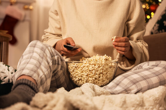 Cozy winter evening at home, person in pajamas eating popcorn and watching television with remote control during holiday season in a warm living room