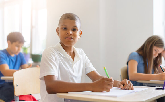 Black schoolboy sitting at his desk with pencil and notebook, looking at camera during classroom lesson while diverse classmates continue studying behind him