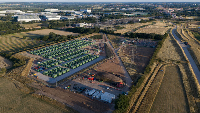Aerial view of a battery energy storage system with rows of green containers, power infrastructure, and surrounding industrial landscape in Hams Hall, England, United Kingdom.