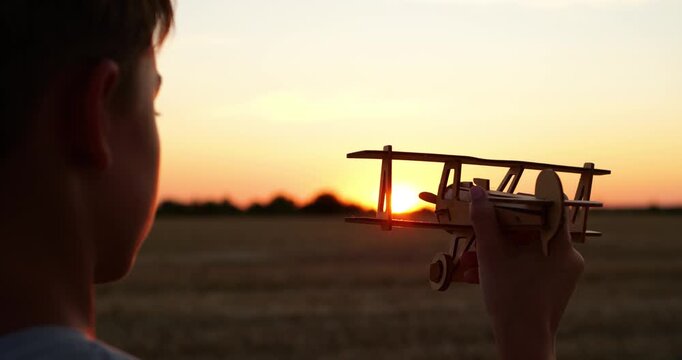 Boy holds vintage wooden biplane toy at sunset in open field. Child imagines flight during golden hour. Nostalgic aviation scene inspires childhood dreams, outdoor play, and creativity.