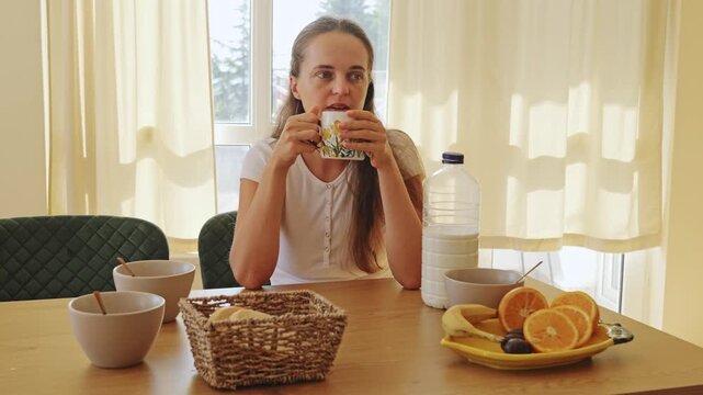 Pensive thoughtful young woman holding a hot ceramic cup sitting alone at the kitchen dining table during peaceful morning breakfast time in a modern apartment