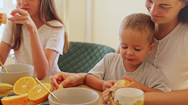 Little toddler boy eating fresh juicy orange slice held gently by mother at the kitchen breakfast table surrounded by family members on a bright sunny day
