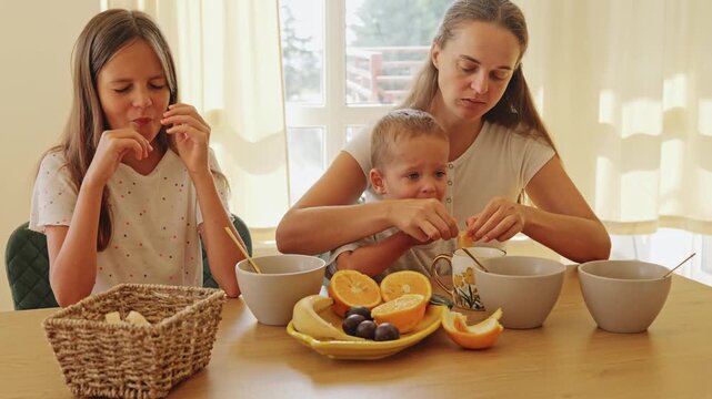 Loving mother sitting with her two children eating fresh orange slices and breakfast at the wooden kitchen table in a cozy home environment during daylight