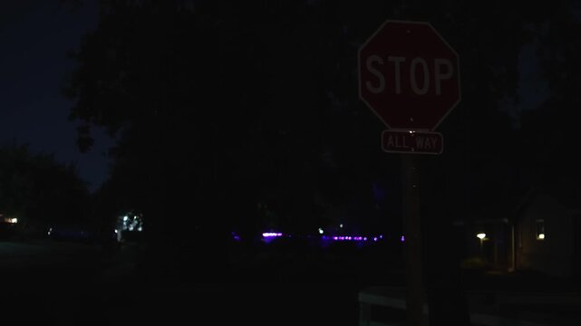 Stop sign at a dark residential neighborhood intersection at night, with an All Way sign against trees, homes and scattered colored lights