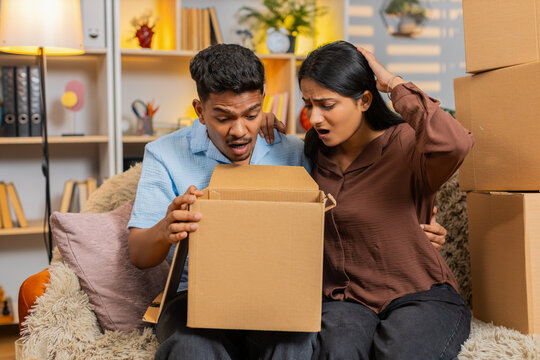 Indian couple man and woman sitting on home sofa open box and react to wrong order with shock. Young family man and woman sitting on home sofa inspect damaged purchase and show shopping frustration.