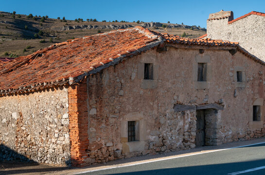 Old stone farmhouse with weathered masonry, small windows, wooden doorway and aged red clay roof tiles stands directly beside a paved rural road. Authentic vernacular architecture illustrates traditio
