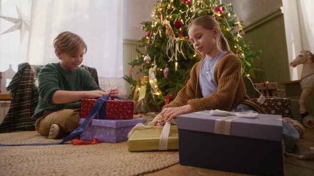 Caucasian boy and girl unwrapping holiday gift boxes sitting near decorated Christmas tree. Perfect for festive seasonal advertising campaigns, retail promotions, and winter family commercials