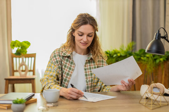 Young woman on home table compares two documents reads terms marks key point and weighs options with focus. Adult girl on home table studies papers carefully before making a final business decision.