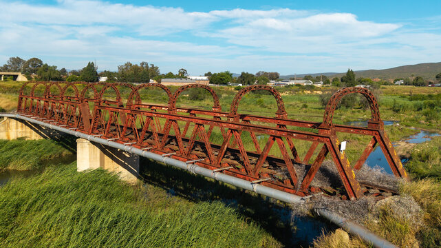 Aerial view of an abandoned rusted railway bridge spanning a river with overgrown vegetation and surrounding rural landscape in Oudtshoorn, Western Cape, South Africa.