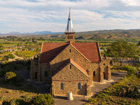 Aerial view of a historic stone church surrounded by arid landscape, native quiver trees, and distant mountains in Oudtshoorn, Western Cape, South Africa.