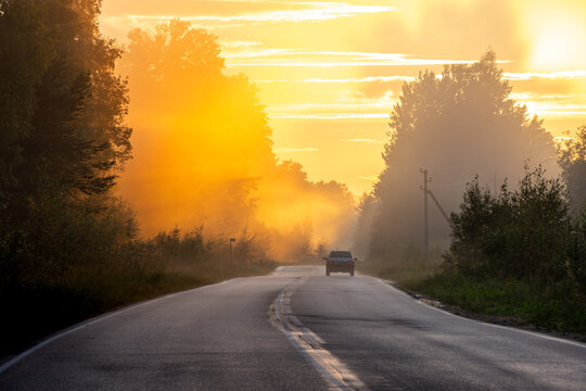 Car driving on empty asphalt road through misty forest into golden sunset sunbeams.