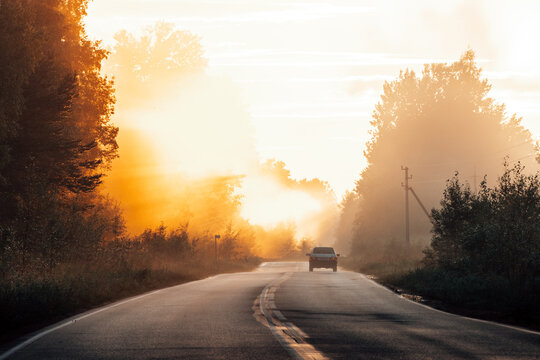 Car driving on empty asphalt road through misty forest into golden sunset sunbeams.