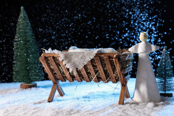 Obraz premium Manger with baby Jesus, angel and Christmas trees on snow against dark background