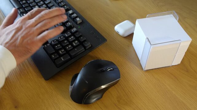 Close-up view of a person using a computer keyboard and mouse at a wooden office desk, with a desktop monitor and wireless earbuds nearby. Modern technology and everyday workspace concept.