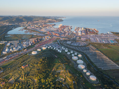 Aerial view of the Sermin industrial area featuring large oil storage tanks, nearby port facilities, and surrounding green hills in Koper, Koper, Slovenia.