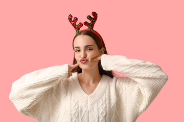 Young woman in sweater with Christmas reindeer horns blowing kiss on pink background © Pixel-Shot