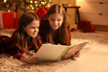 Obraz premium Cute happy little girls in checkered pajamas with book reading Christmas story at night