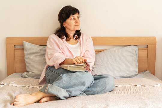 Mature woman sits cross-legged on bed, closed book and glasses rest on her lap. She is wearing pink shirt and jeans, set against backdrop of a bright bedroom. Slow living, quiet unhurried morning