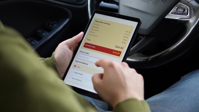 Shopper reviews an online sale checkout on a tablet, comparing discounts, promo savings and payment options during a digital retail purchase inside a car.