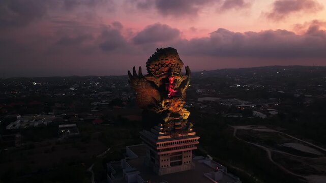 Garuda Wisnu Kencana sunset view reveals the illuminated Hindu monument rising above southern Bali with detailed sculpture and dramatic dusk twilight horizon orbit aerial drone footage