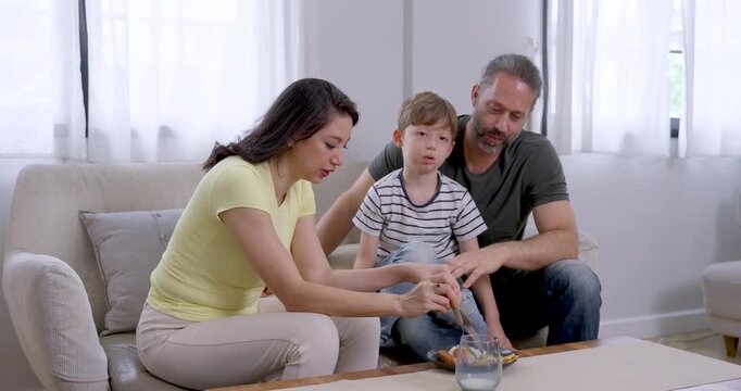 Asian mother serves food from bowl while Caucasian father guides young son, who lifts pizza slice and eats on sofa, creating attentive parenting, shared snack time, and warm family bonding at home.